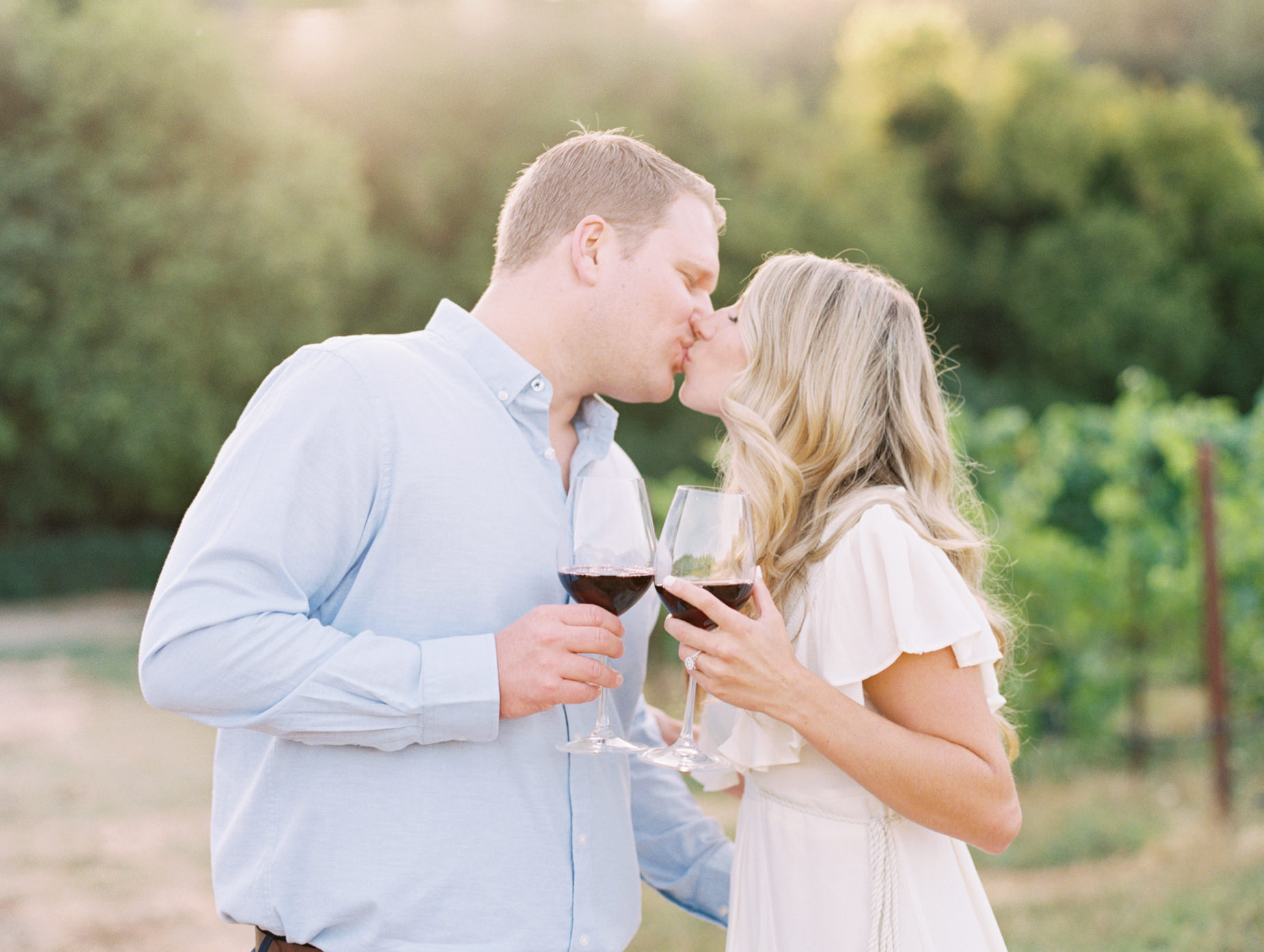 Jeff and Heather kiss while holding wine glasses at vineyard engagement at Williams Selyem Wines
